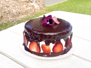 Chocolate cake with fresh strawberries and cream between layers, topped with glossy chocolate ganache and edible flowers, displayed on a white outdoor table.
