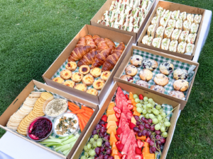 A selection of freshly made catering boxes featuring sandwiches, wraps, pastries, muffins, fruit and a grazing platter, displayed on a table outdoors.