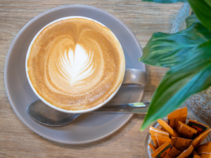 Freshly made latte with latte art served in a grey cup and saucer on a timber table at STEPS Café on George, with a spoon, sugar sachets, and greenery nearby.