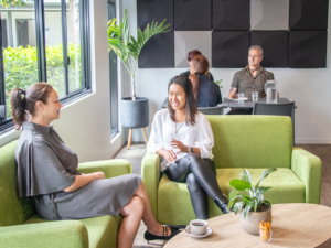 Customers seated in a bright, modern café lounge with green armchairs, timber tables, indoor plants and large windows providing natural light.