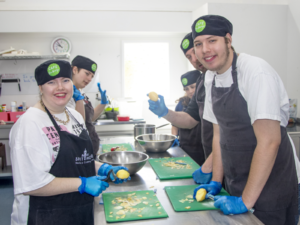STEPS Café on George staff and participants preparing food in a commercial kitchen, wearing aprons and gloves while peeling and chopping ingredients.