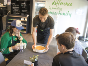 A café team member serves a bowl of soup to diners seated at a table inside STEPS Café on George.